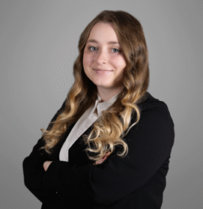 Young female attorney with long wavy blonde hair, arms crossed, smiling softly in black blazer and white shirt against gray background.