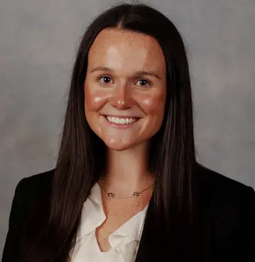 Young female attorney with long wavy blonde hair, arms crossed, smiling softly in black blazer and white shirt against gray background.